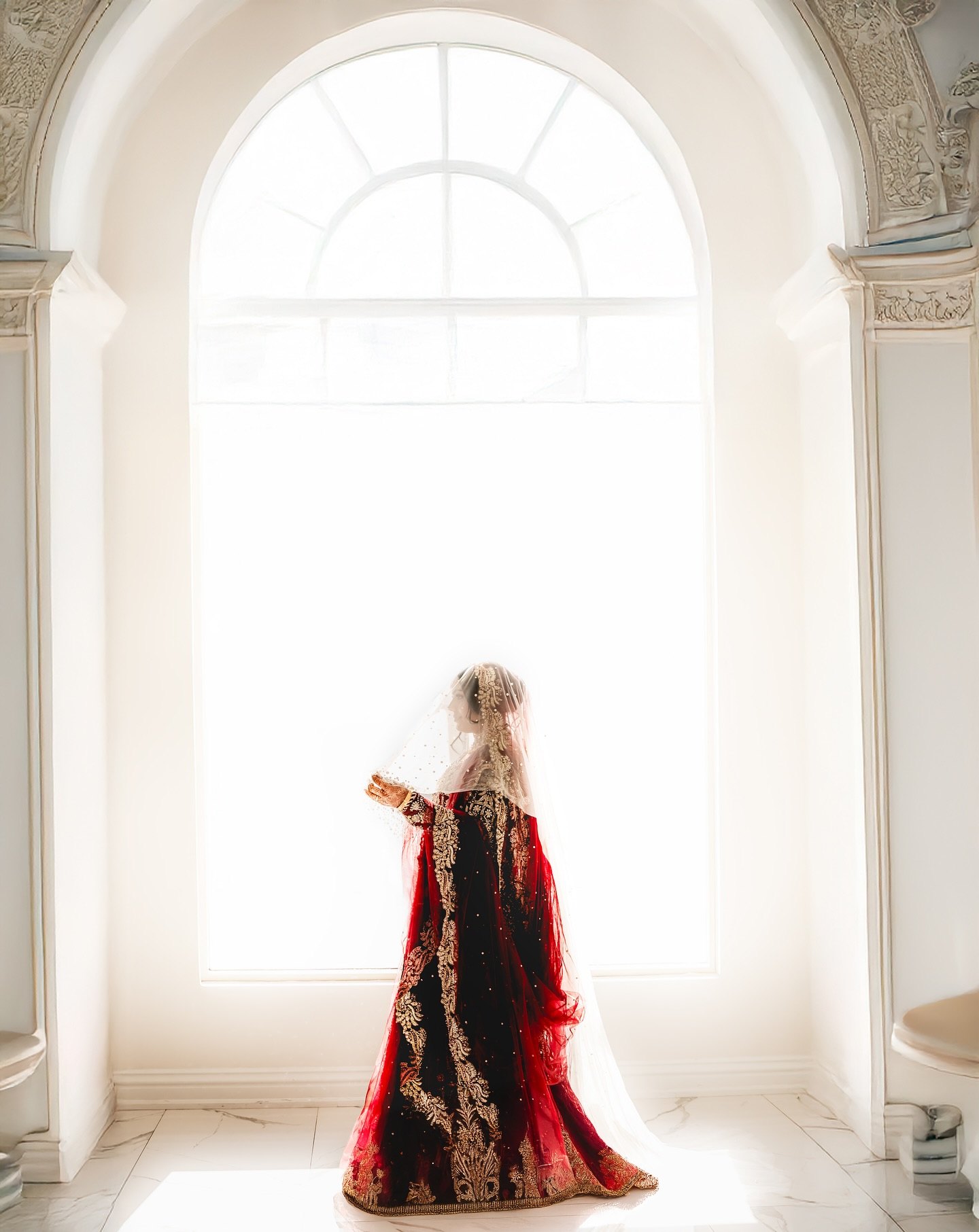 The bride in a deep red embroidered shawl stands before a tall arched window full of light