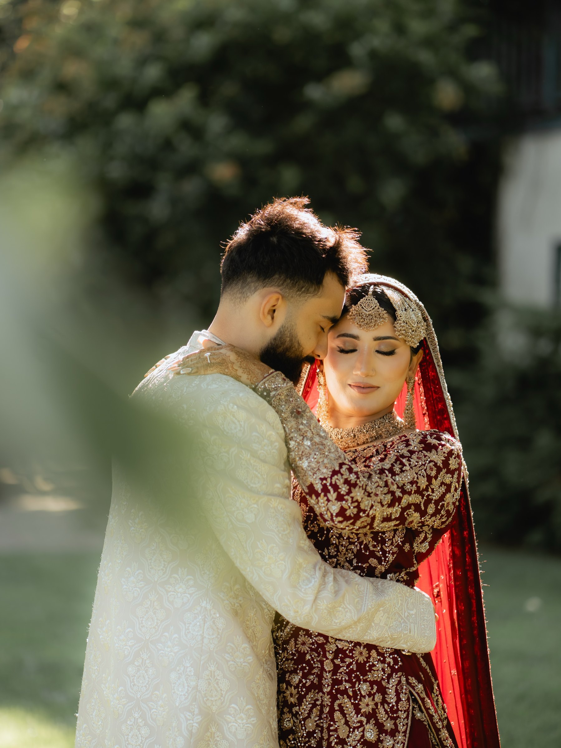 The groom in an ivory sherwani holds the bride in a maroon and gold lehenga in a sunlit garden