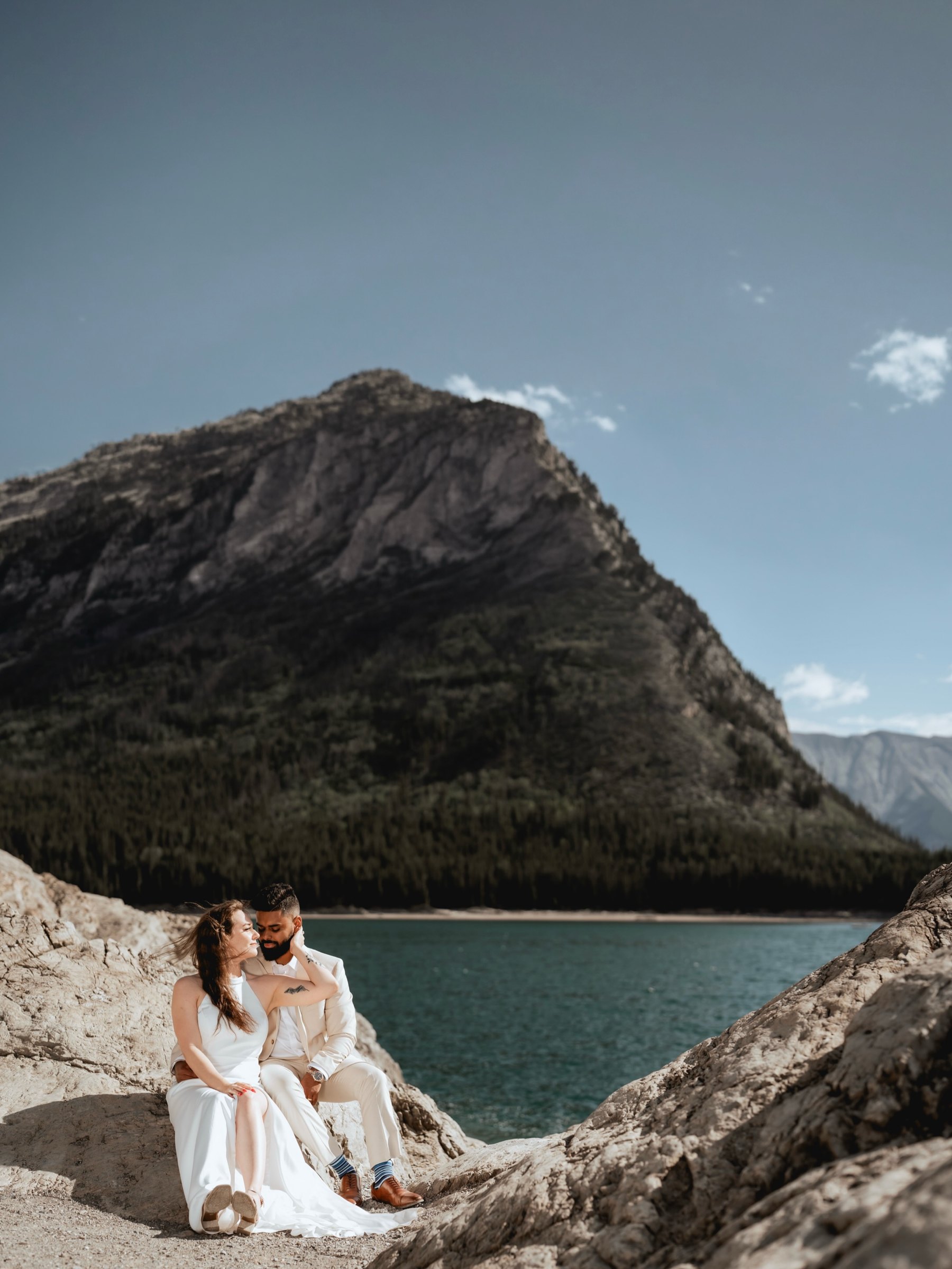 An engaged couple sit on rocks above a mountain lake beneath a grey peak
