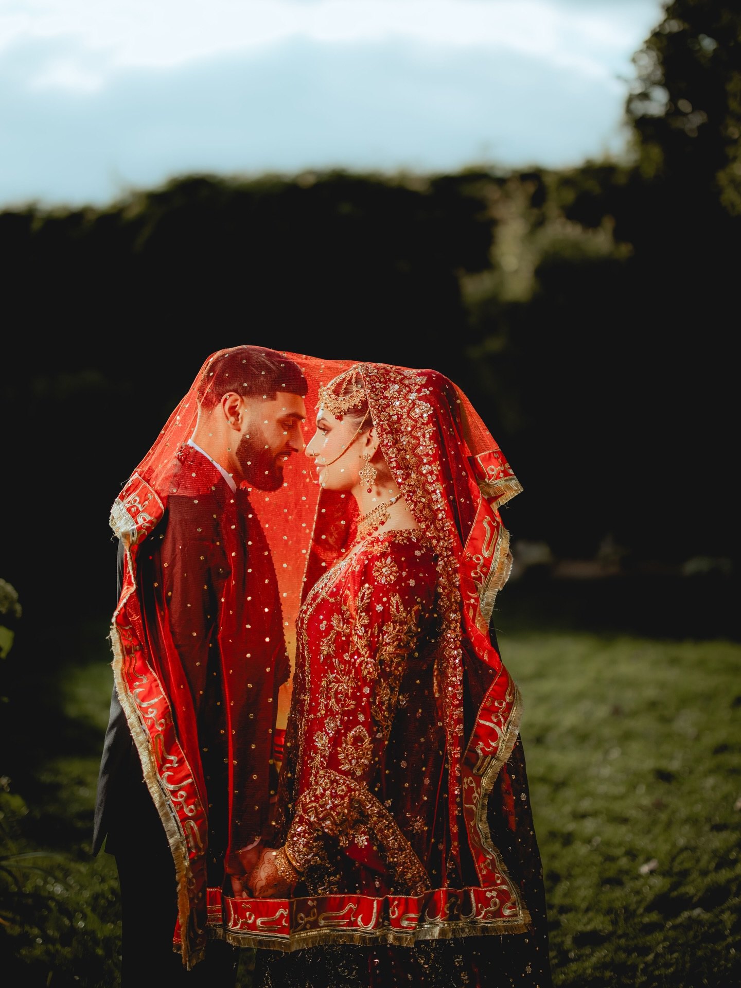 Bride and groom sheltered together beneath her red embroidered dupatta at golden hour