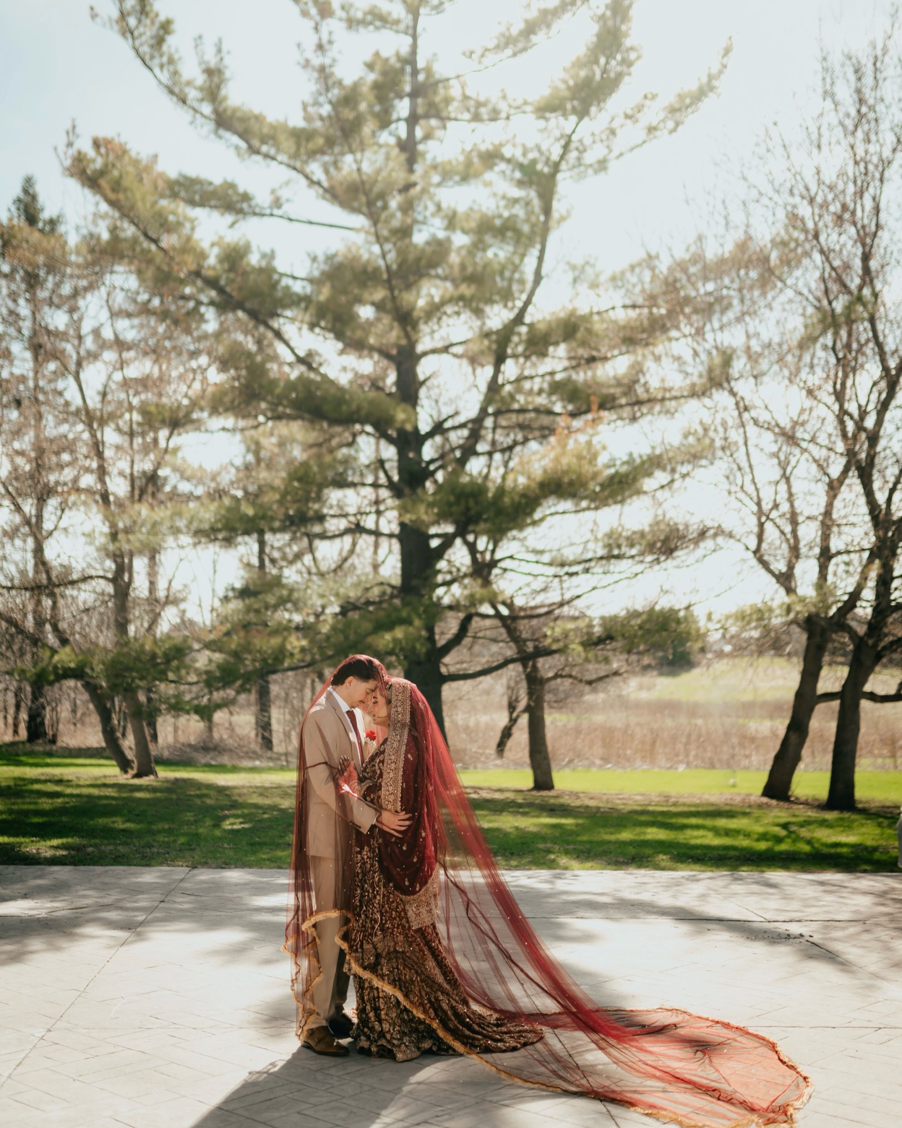 The groom kisses the bride beneath tall pines, her long red dupatta trailing across the stone